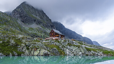 Eine Hütten in den Bergen, im Vordergrund ein kleiner Bergsee.