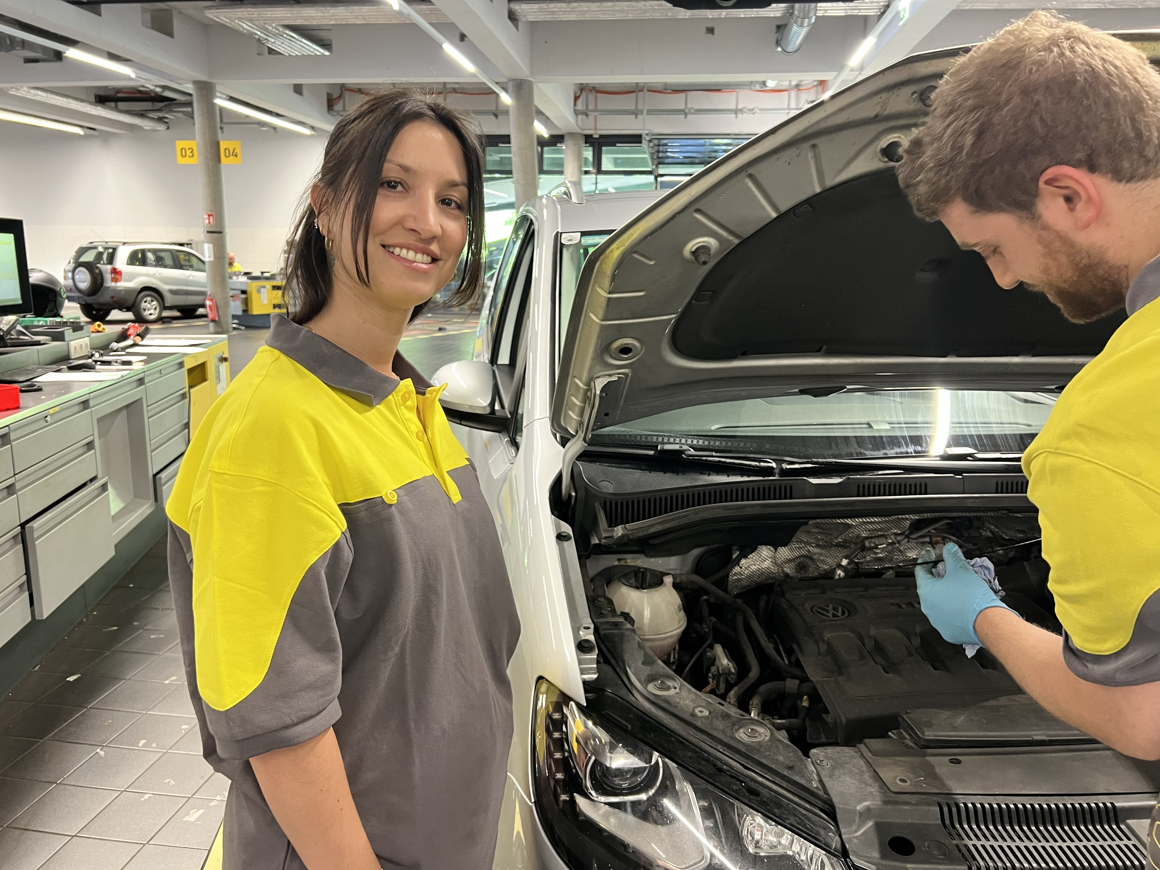 Auf dem Bild ist eine Frau zu sehen, die in die Kamera lacht und vor einem Auto mit offener Motorhaube steht. Sie befindet sich in einer Werkstatt. Neben ihr steht ein Mann, der dem Auto zugewandt ist und gerade etwas repariert.