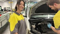 Auf dem Bild ist eine Frau zu sehen, die in die Kamera lacht und vor einem Auto mit offener Motorhaube steht. Sie befindet sich in einer Werkstatt. Neben ihr steht ein Mann, der dem Auto zugewandt ist und gerade etwas repariert.