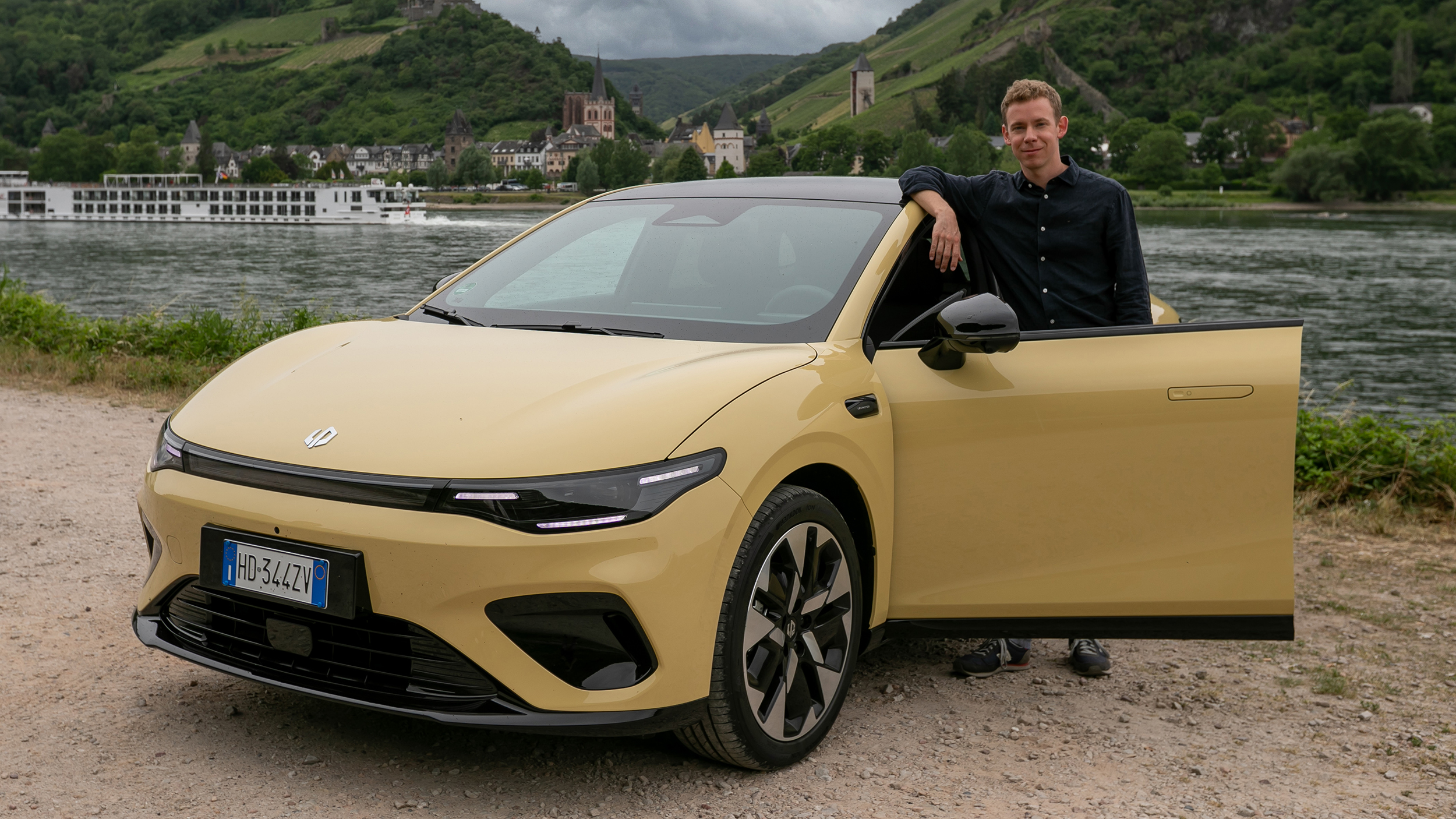 Ein Mann in dunklem Hemd lehnt hinter der geöffneten Fahrertür an einem gelben Auto.
