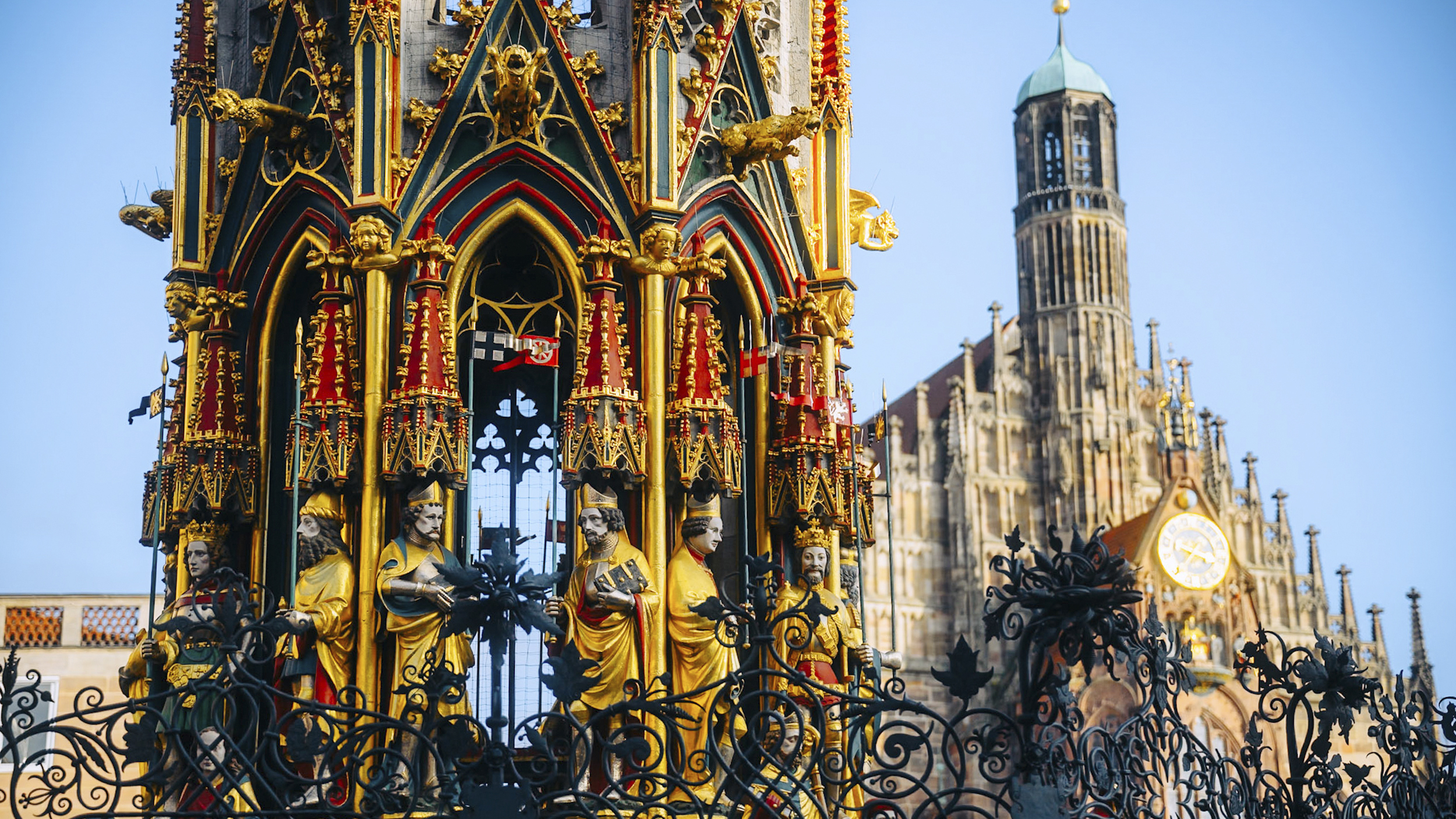 Ein bunter Brunnen im gotischen Stil, im Hintergrund eine Kirche.