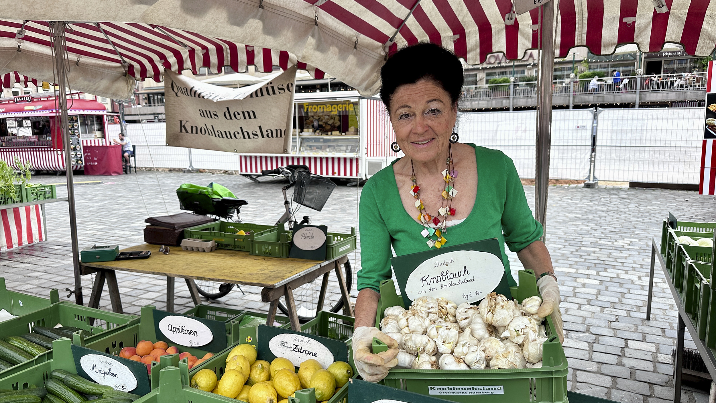 Eine Marktstandlerin im grünen Gewand präsentiert stolz eine Kiste Knoblauch.