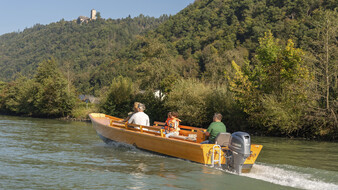 Zille fährt auf der Donau mit 3 Personen darin, im Hintergrund bewaldete Uferlandschaft.