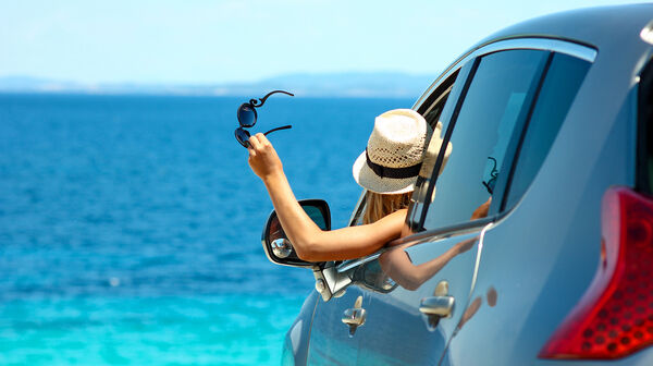 Eine Frau mit Strohhut hält ihre Sonnenbrille aus dem Autofenster und blickt aufs Meer.
