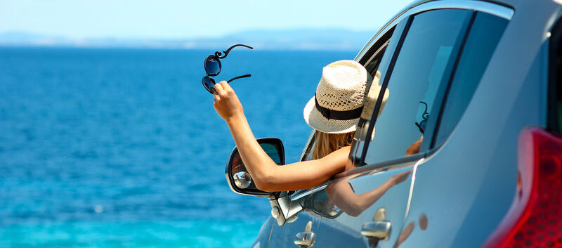 Eine Frau mit Strohhut hält ihre Sonnenbrille aus dem Autofenster und blickt aufs Meer.