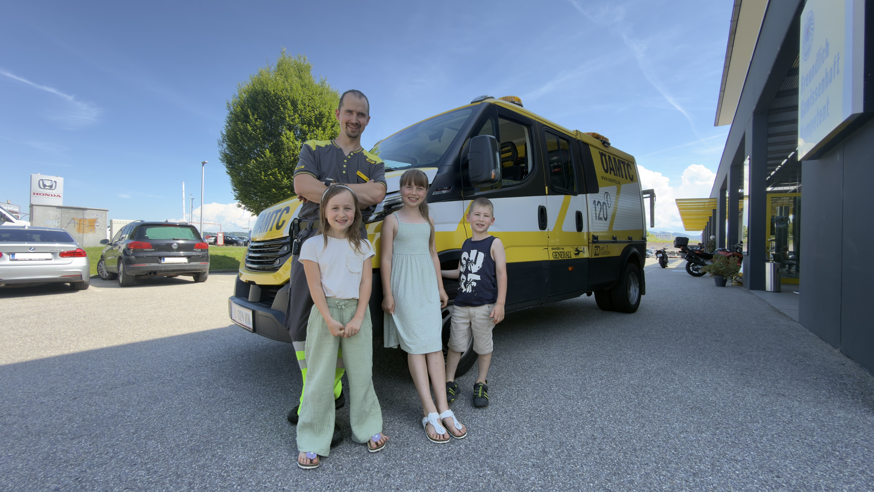 Es ist ein Mann mit drei Kindern zu sehen, die vor einem ÖAMTC Pannenfahrzeug stehen.