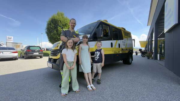 Es ist ein Mann mit drei Kindern zu sehen, die vor einem ÖAMTC Pannenfahrzeug stehen.