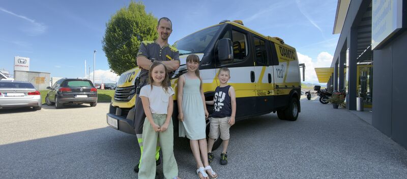 Es ist ein Mann mit drei Kindern zu sehen, die vor einem ÖAMTC Pannenfahrzeug stehen.