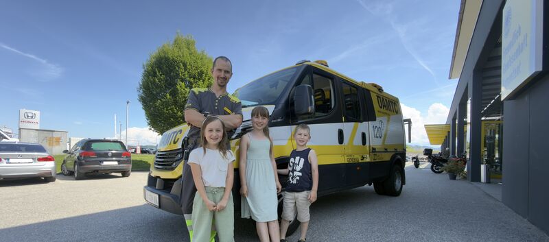 Es ist ein Mann mit drei Kindern zu sehen, die vor einem ÖAMTC Pannenfahrzeug stehen.