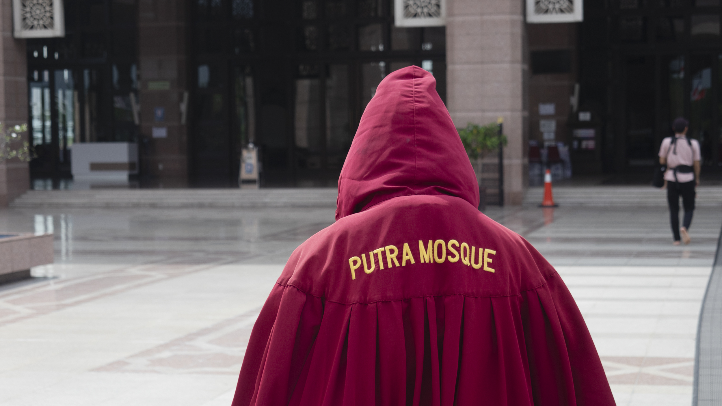 Eine Person geht mit einem roten Ganzkörper-Kleid, auf dem hinten "Putra Mosque" steht, durch die Putra-Moschee.
