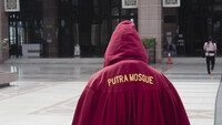 Eine Person geht mit einem roten Ganzkörper-Kleid, auf dem hinten "Putra Mosque" steht, durch die Putra-Moschee.