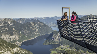 Zwei Wanderer stehen auf einer Aussichtsplattform, Blick auf das Dachsteinmassiv mit See.