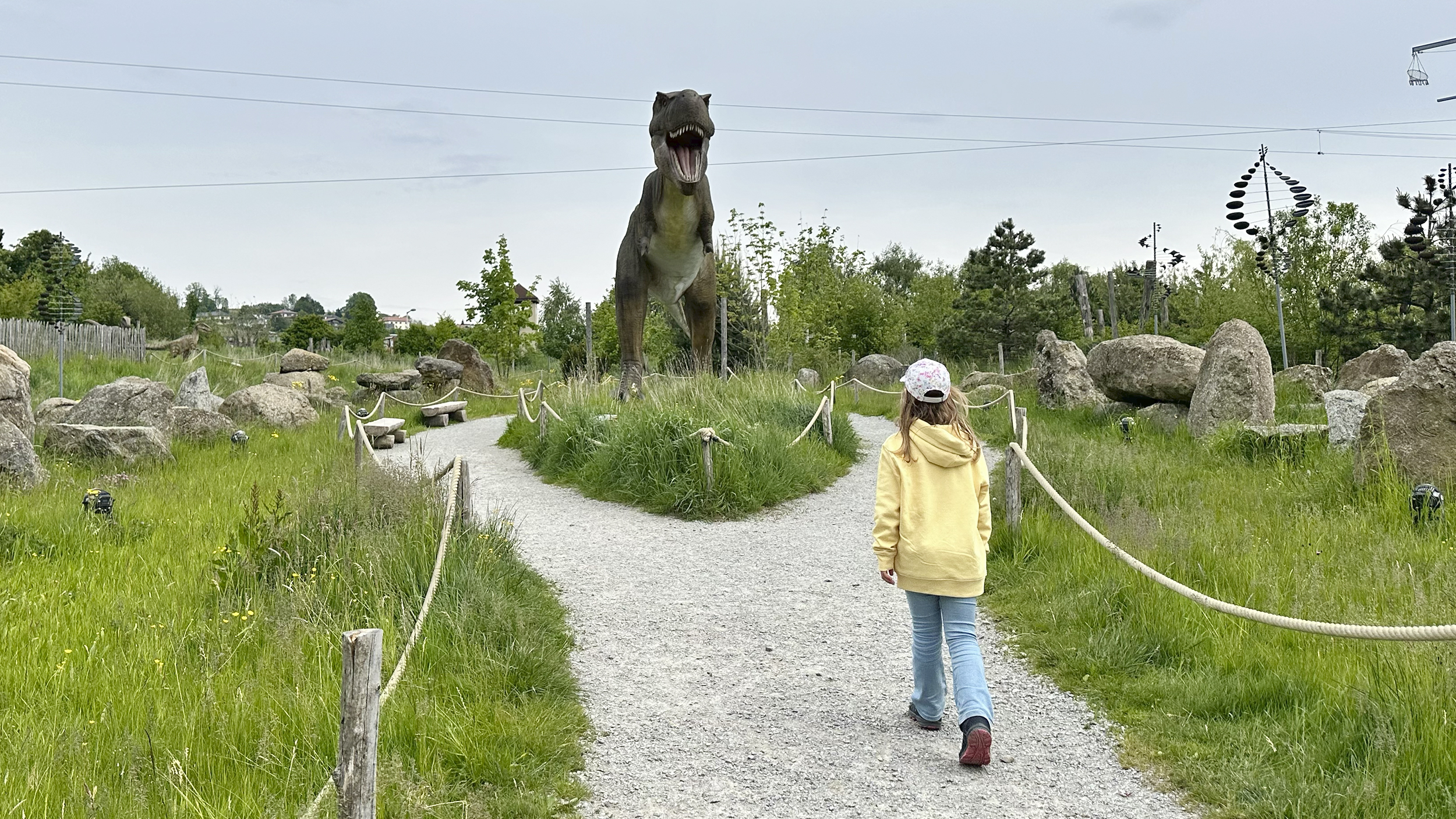 Ein Mädchen im gelben Hoodie geht auf einem Weg auf eine große Dinosaurierstatue zu.