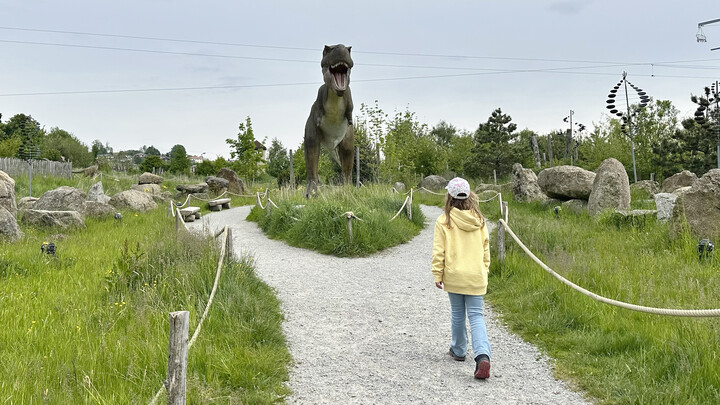 Ein Mädchen im gelben Hoodie geht auf einem Weg auf eine große Dinosaurierstatue zu.