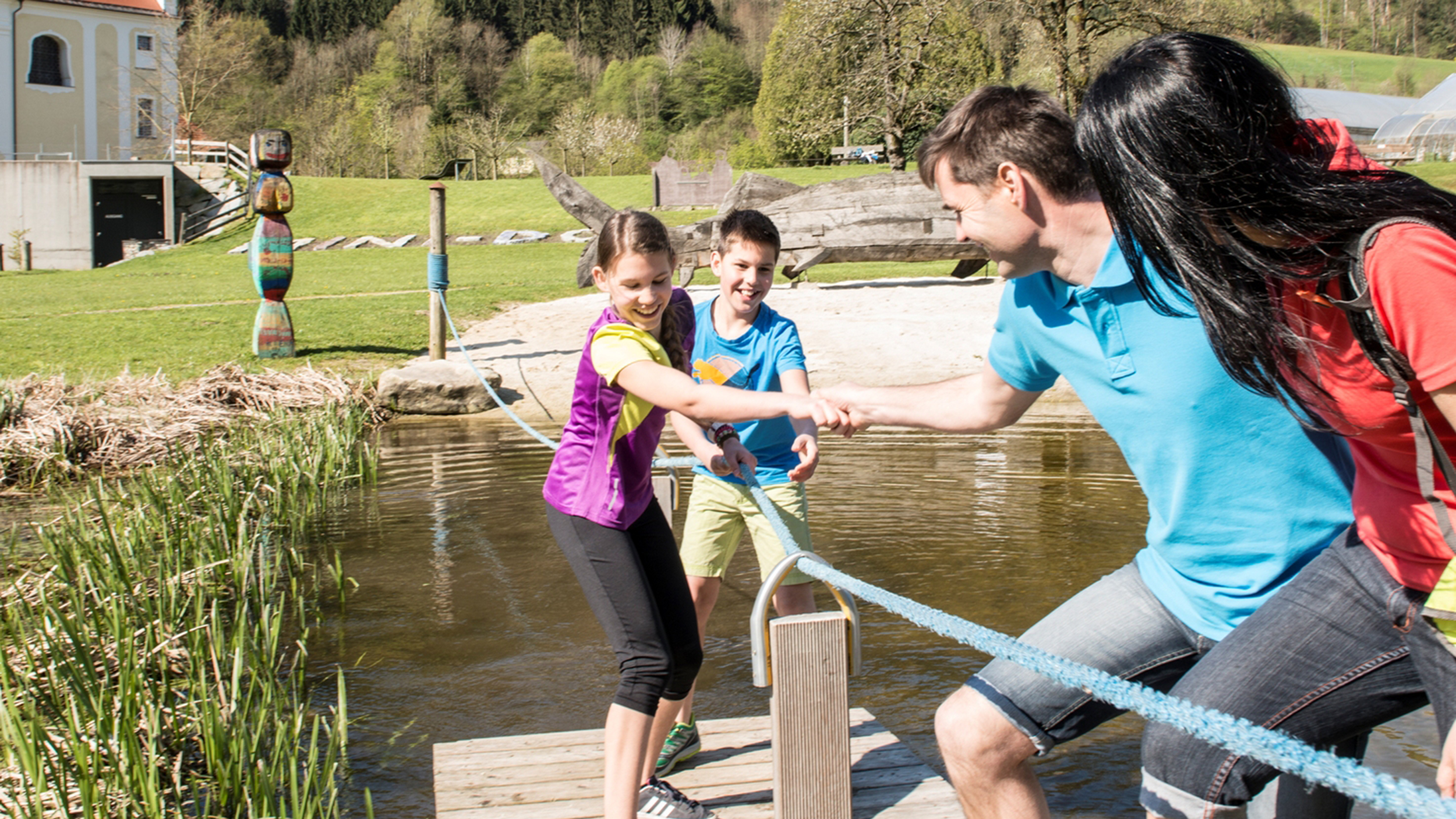 4 Kinder spielen in der Engelhartszeller Donau-Welt auf einem Floß am Wasser und ziehen sich mit einem Seil zum Ufer.