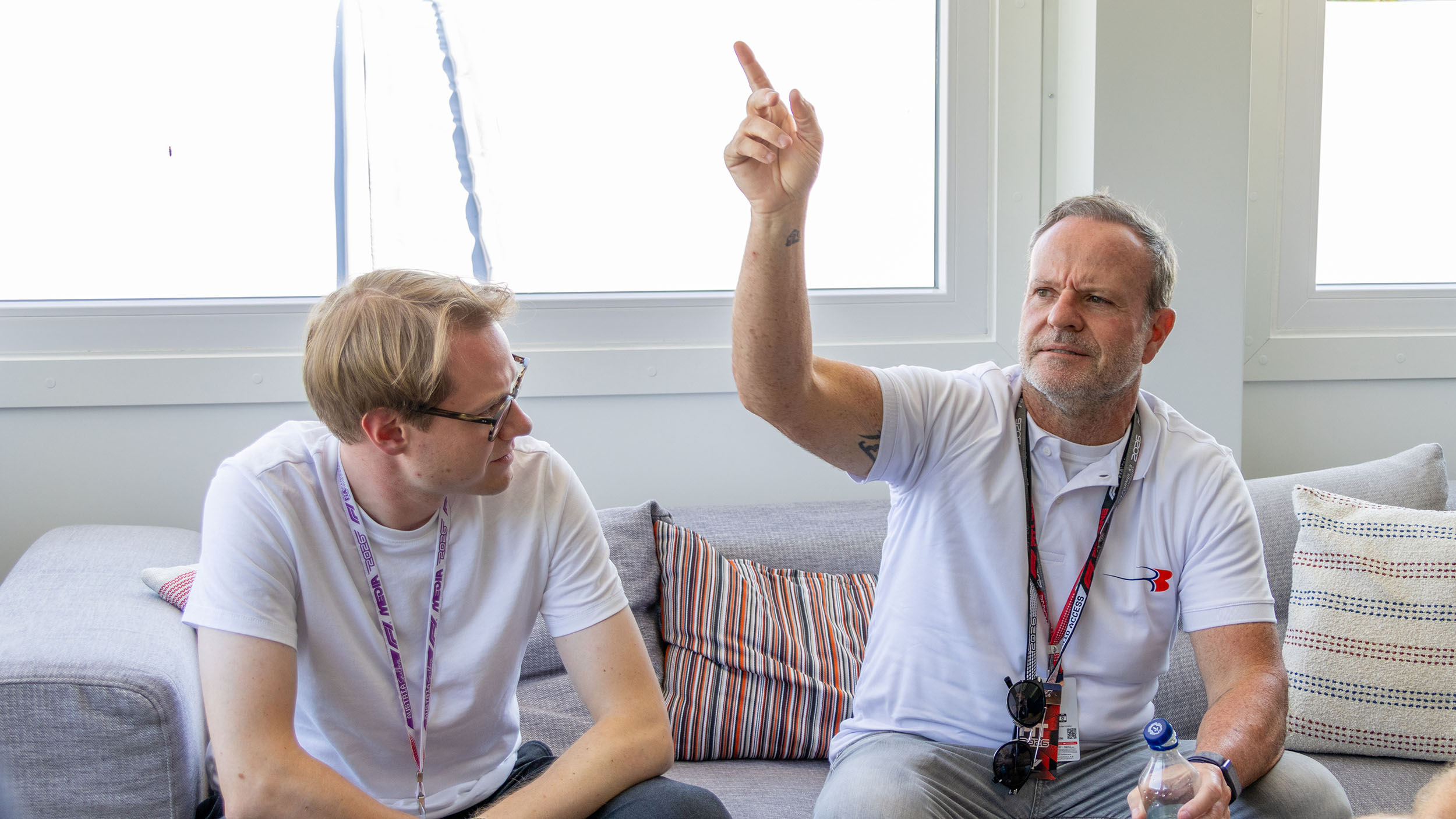 Rubens Barrichello sitzt auf einer Couch mit einem Journalisten und zeigt mit der Hand in die Höhe.