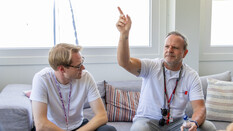 Rubens Barrichello sitzt auf einer Couch mit einem Journalisten und zeigt mit der Hand in die Höhe.