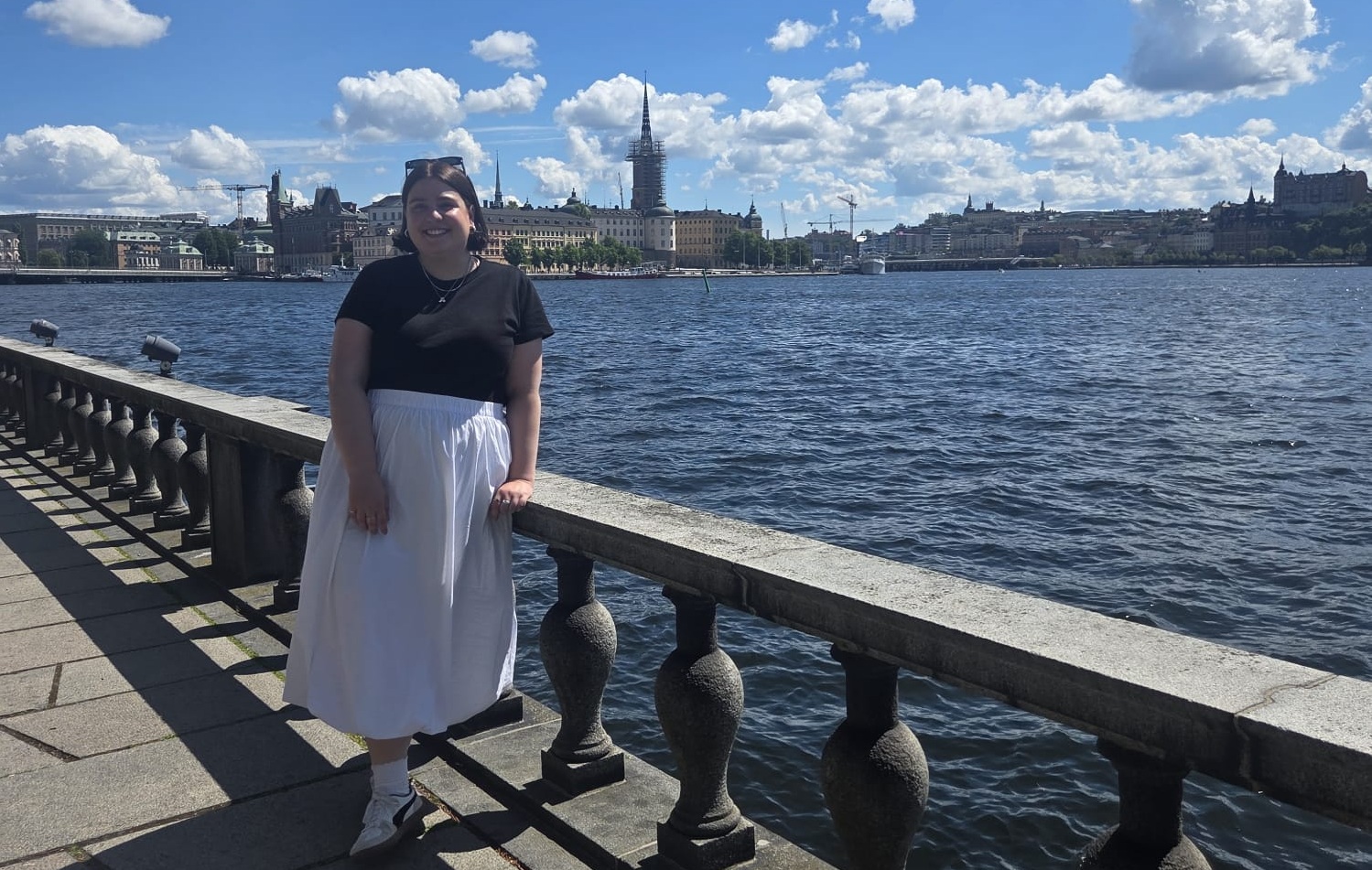 Auf dem Foto ist eine Frau im weißen Rock und schwarzem Tshirt zu sehen. Sie lehnt an einem Geländer. Hinter ihr sieht man das Meer und die Altstadt von Stockholm.