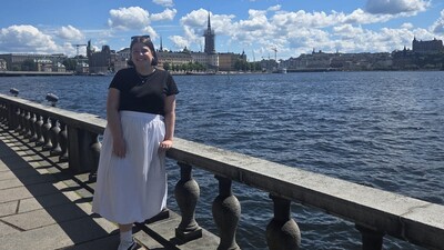 Auf dem Foto ist eine Frau im weißen Rock und schwarzem Tshirt zu sehen. Sie lehnt an einem Geländer. Hinter ihr sieht man das Meer und die Altstadt von Stockholm.