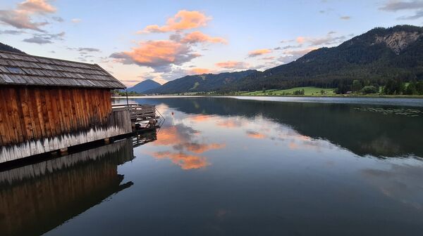 Das Foto zeigt den Weissensee, auf der linken Seite sieht man ein Holzhaus und recht einen Berg.