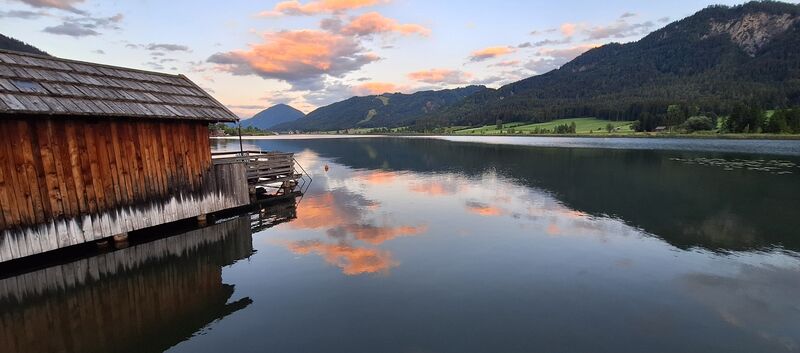 Das Foto zeigt den Weissensee, auf der linken Seite sieht man ein Holzhaus und recht einen Berg.