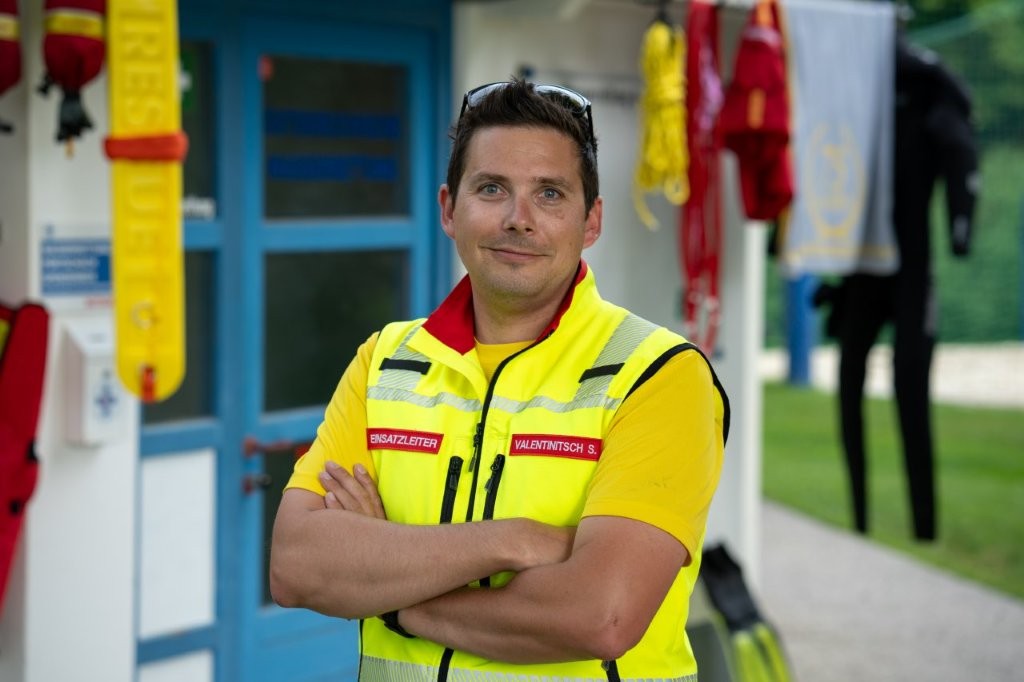 Auf dem Bild sieht man einen Mitarbeiter der Wasserrettung. Er steht mit verschränkten Armen und leuchtgelber Einsatzbekleidung vor einer Hütte.