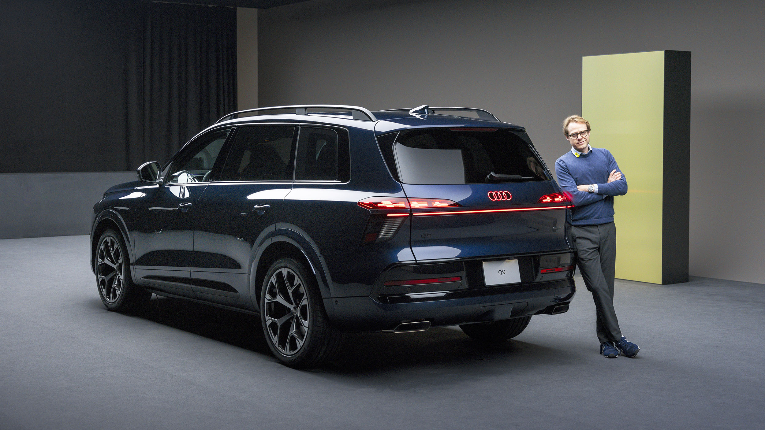 Eine Person lehnt am Heck eines blauen Audi Q9, der in einem Studio steht.