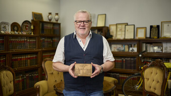 ÖAMTC-Archivar Joachim Moser, trägt ein weißes Kurzarmhemd mit Pullunder. Er steht im Archiv, umgeben von Büchern und hält eine Plakette.