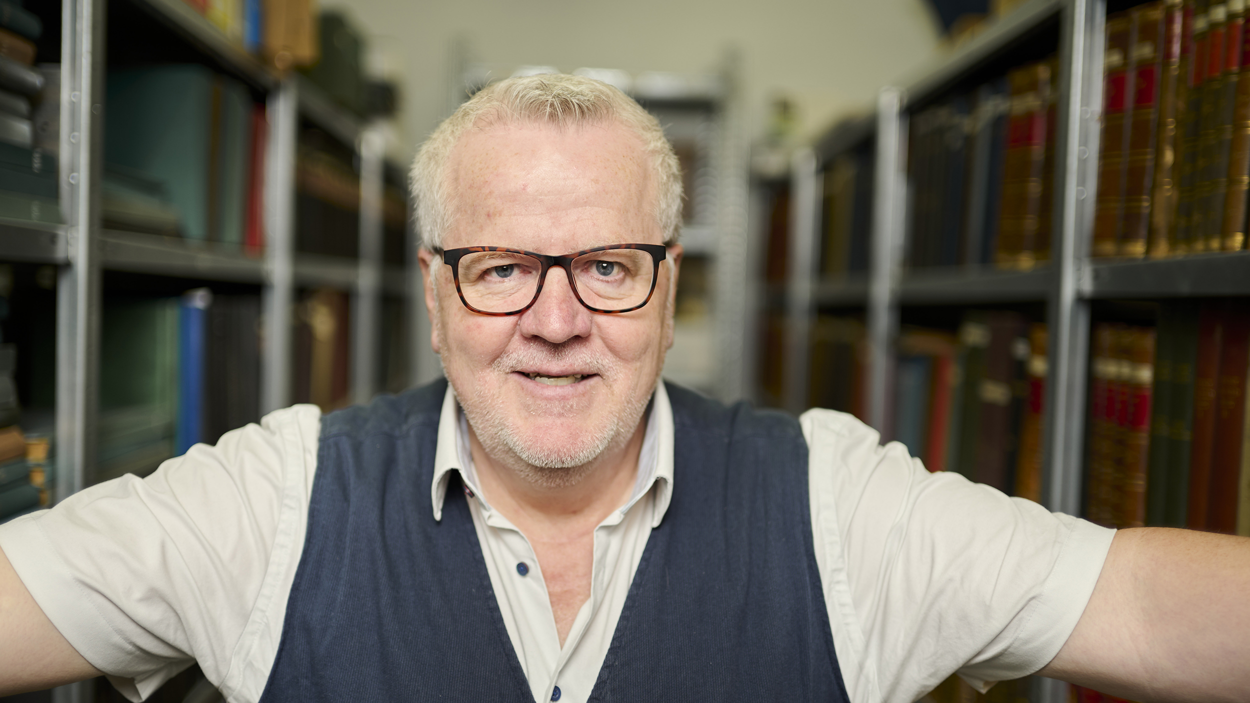 ÖAMTC-Archivar Joachim Moser, trägt ein weißes Kurzarmhemd mit Pullunder. Er steht im Archiv, umgeben von Büchern.