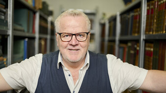 ÖAMTC-Archivar Joachim Moser, trägt ein weißes Kurzarmhemd mit Pullunder. Er steht im Archiv, umgeben von Büchern.