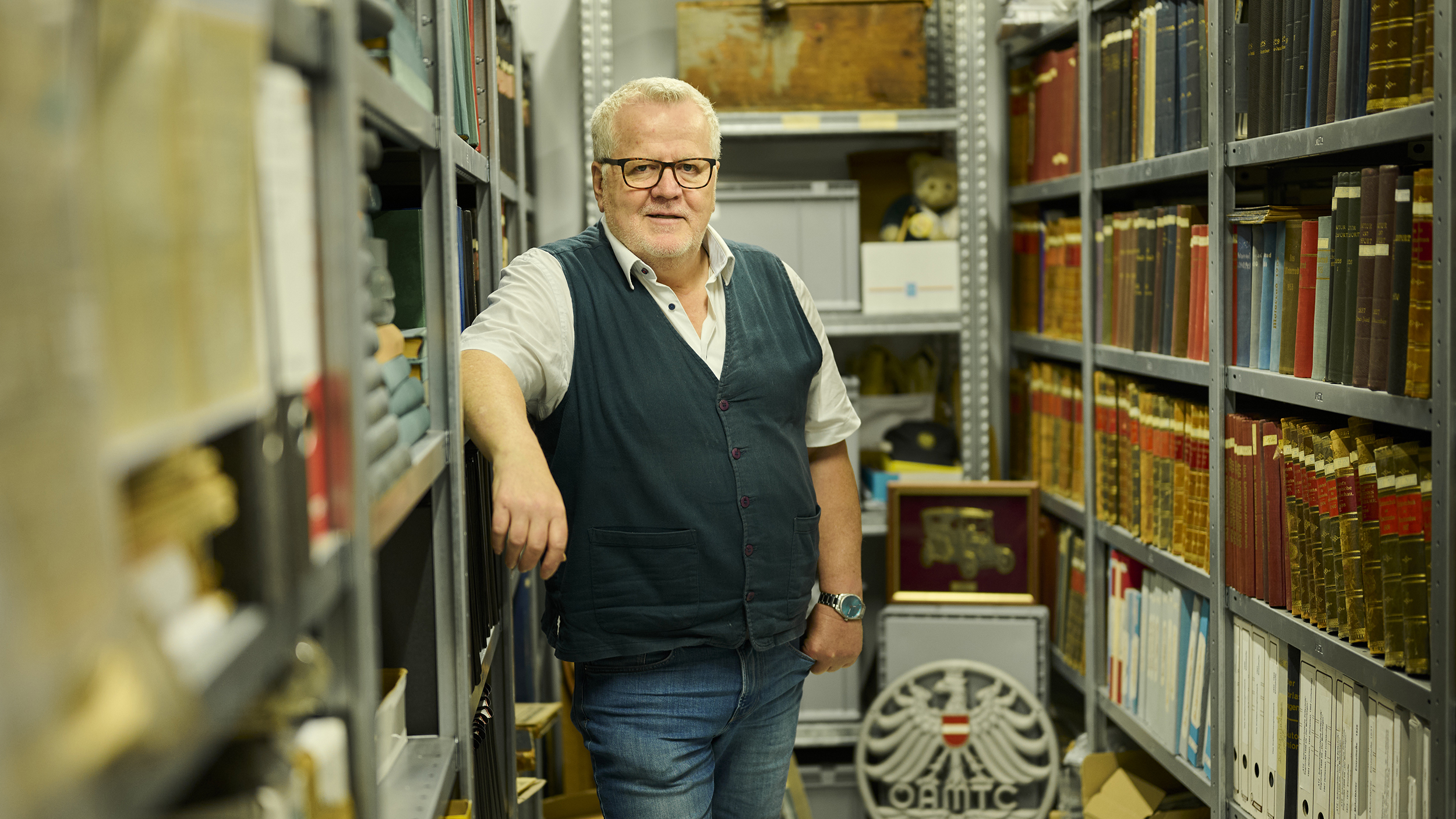ÖAMTC-Archivar Joachim Moser, trägt ein weißes Kurzarmhemd mit Pullunder. Er steht im Archiv, umgeben von Büchern.