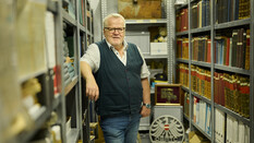 ÖAMTC-Archivar Joachim Moser, trägt ein weißes Kurzarmhemd mit Pullunder. Er steht im Archiv, umgeben von Büchern.
