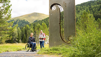 Eine Rollstuhlfahrer und Begleitung auf einem Wanderweg zur Nockalm neben einer Skulptur.