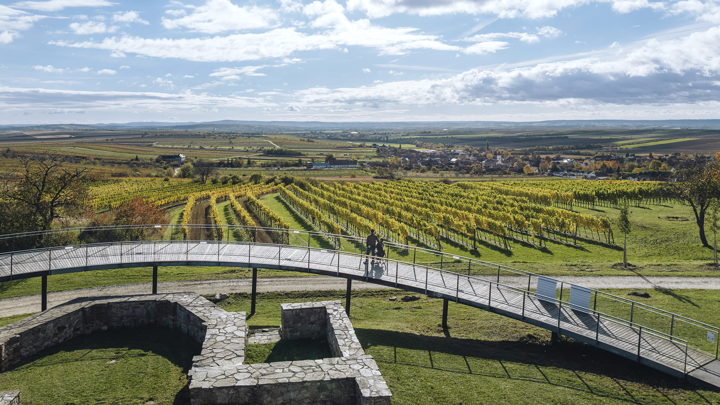 Eine Ausblicksrampe bietet weiten Blick über Weingärten im Weinviertel. Eine Person im Rollstuhl samt Begleitung genießen den Ausblick. 