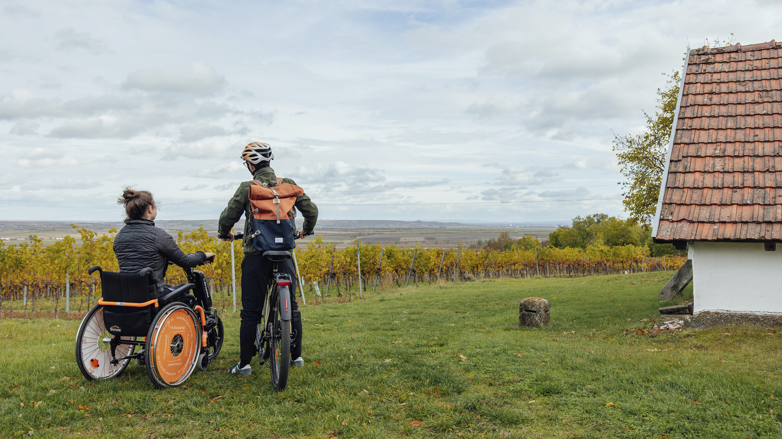 Eine Rollstuhlfahrerin im E-Rollstuhlbike steht neben einem Radfahrer, beide blicken auf Weingärten neben einem kleinen Haus.