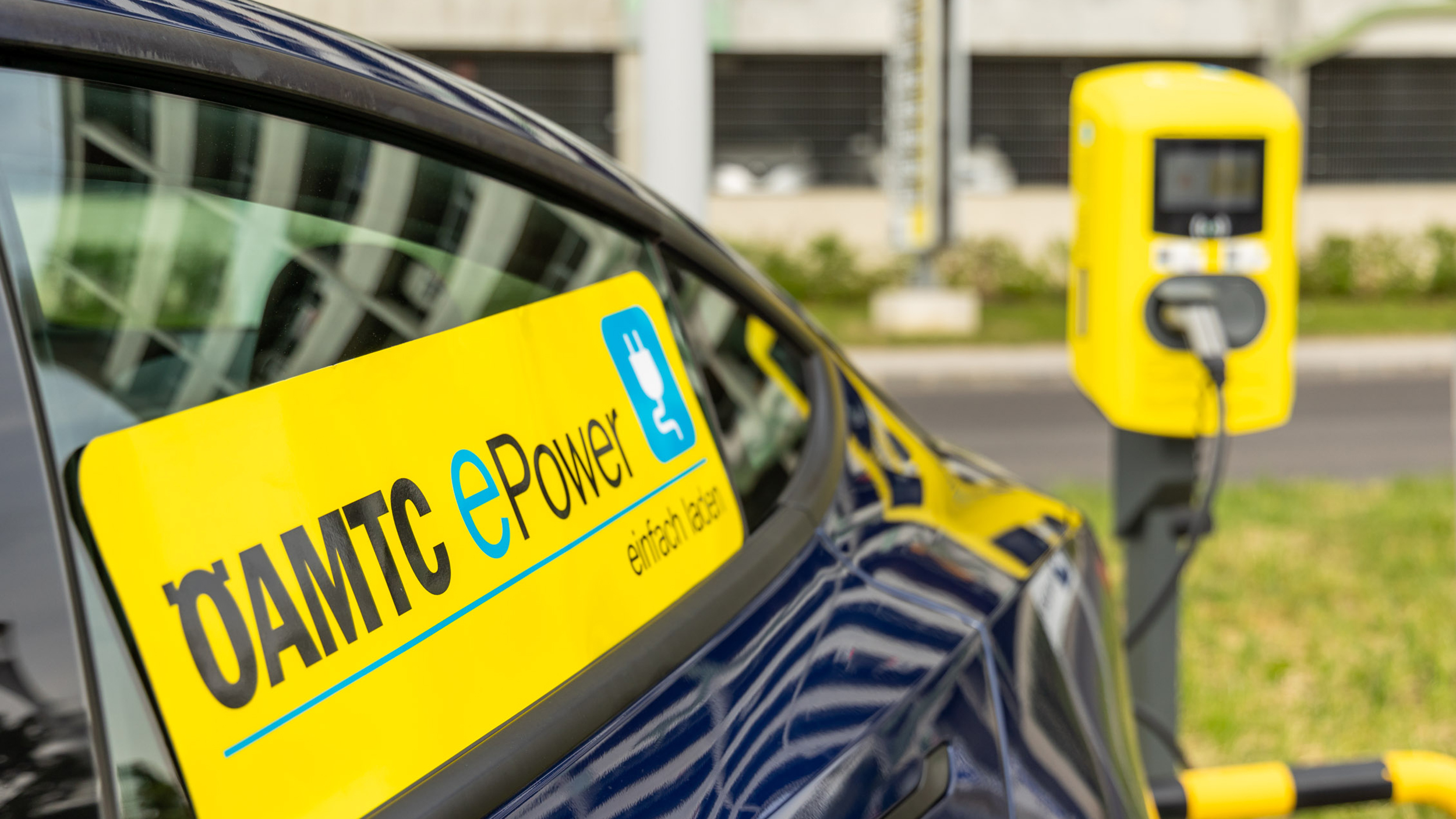 Ein gelber ÖAMTC ePower-Sticker auf hinteren Fensterscheibe eines Autos an einer Ladesäule.