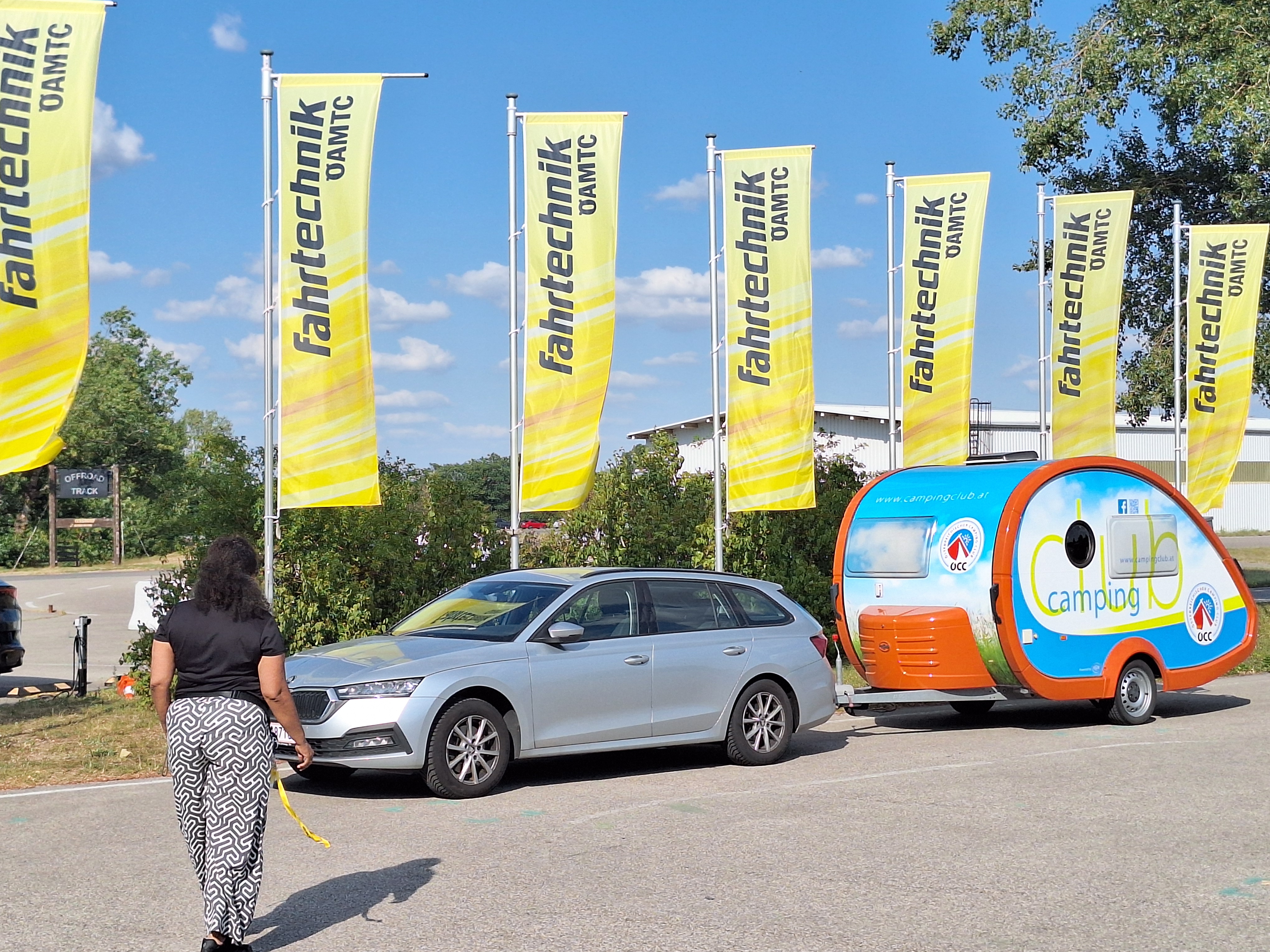 Es ist eine Frau von hinten zu sehen, die auf ein silbernes Auto mit Camping-Anhänger zugeht. Hinter dem Auto wehen gelbe Fahnen.