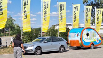 Es ist eine Frau von hinten zu sehen, die auf ein silbernes Auto mit Camping-Anhänger zugeht. Hinter dem Auto wehen gelbe Fahnen.
