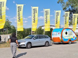 Es ist eine Frau von hinten zu sehen, die auf ein silbernes Auto mit Camping-Anhänger zugeht. Hinter dem Auto wehen gelbe Fahnen.