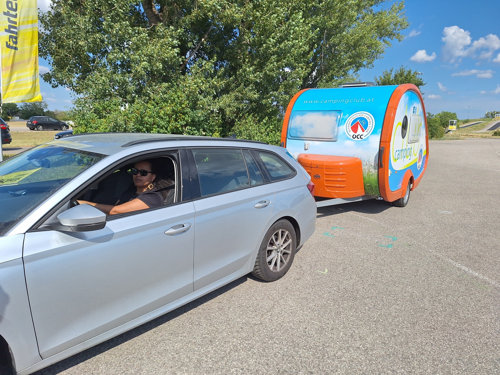 Man sieht eine Frau, die in einem grauen Auto mit Camping-Anhänger sitzt.