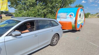 Man sieht eine Frau, die in einem grauen Auto mit Camping-Anhänger sitzt.