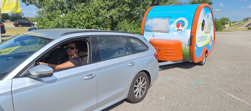 Man sieht eine Frau, die in einem grauen Auto mit Camping-Anhänger sitzt.
