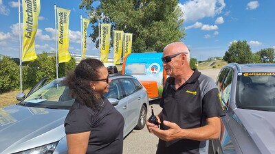 Man sieht eine Frau und einen Mann, die sich unterhalten. Im Hintergrund sind gelbe Fahnen und Autos.