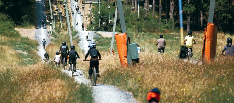 Mountainbiker:innen fahren neben einem Schlepplift einen sommerlichen Berghang hinauf.