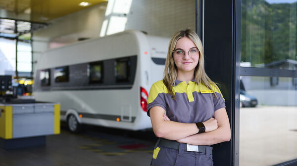 Eine junge KFZ-Technikerin steht an eine Tür gelehnt vor der ÖAMTC-Werkstatt. Im Hintergrund steht ein Wohnwagen.