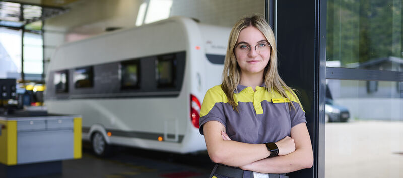Eine junge KFZ-Technikerin steht an eine Tür gelehnt vor der ÖAMTC-Werkstatt. Im Hintergrund steht ein Wohnwagen.