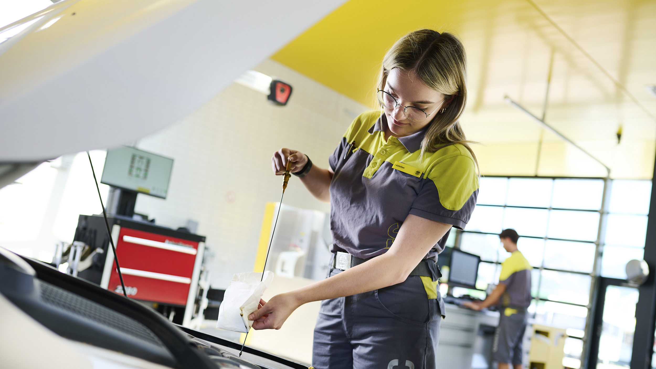Jessica, ein junger Lehrling in Ausbildung zur Kfz-Technikerin steht vor geöffneter Motorhaube in der ÖAMTC-Werkstatt und kontrolliert den Ölpegel.
