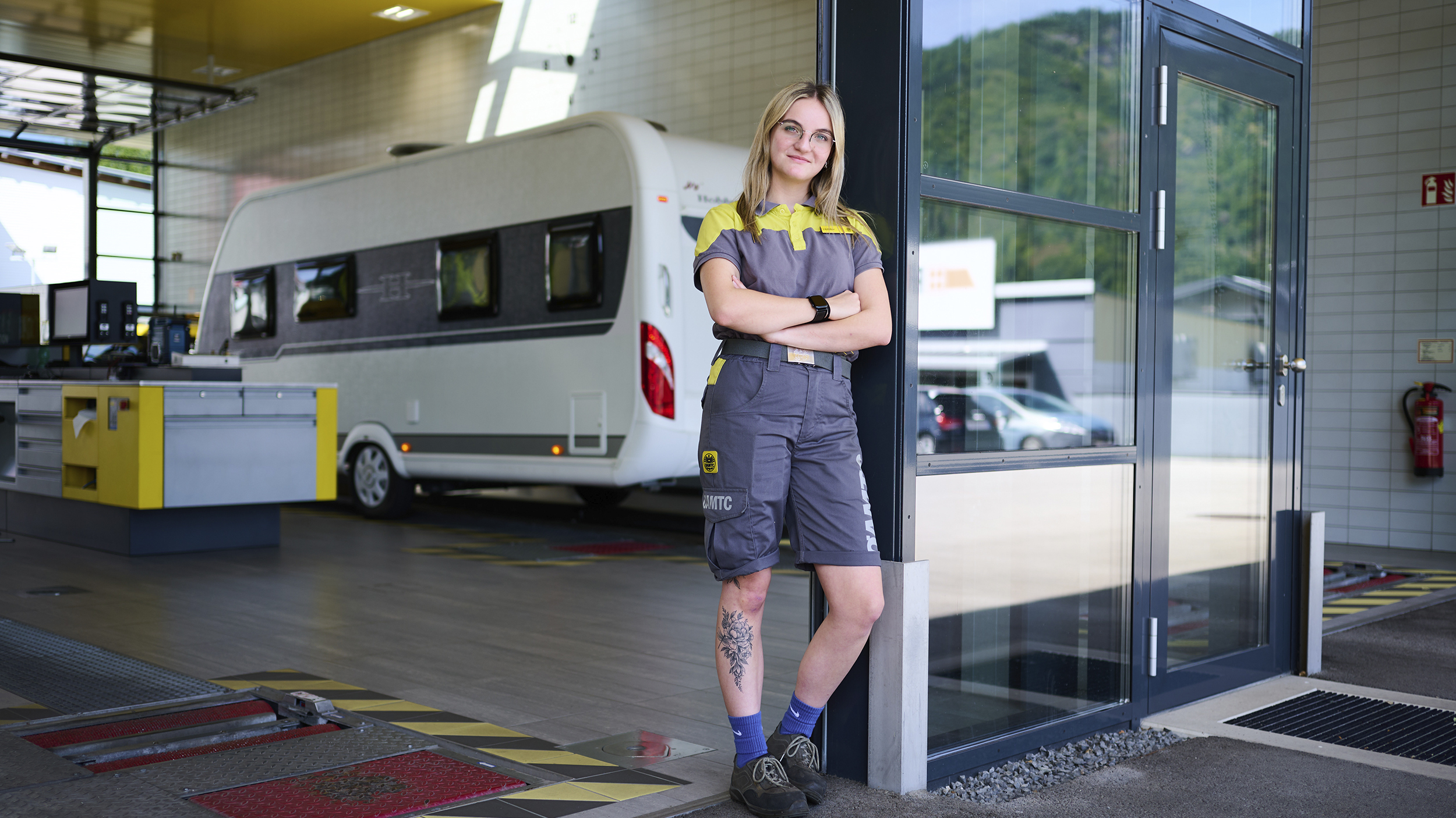 Eine junge KFZ-Technikerin steht an eine Tür gelehnt vor der ÖAMTC-Werkstatt. Im Hintergrund steht ein Wohnwagen.