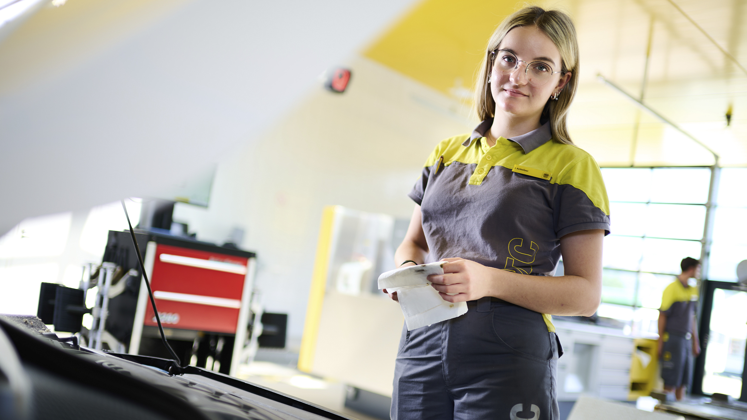 Jessica, ein junger Lehrling in Ausbildung zur Kfz-Technikerin steht vor geöffneter Motorhaube in der ÖAMTC-Werkstatt und kontrolliert den Ölpegel.
