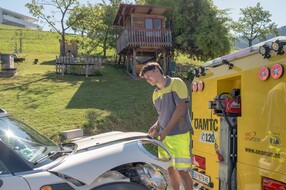 Auf dem Bild sieht man einen jungen Mitarbeiter des ÖAMTC. er öffnet gerade die Motorhaube eines weißen Autos.